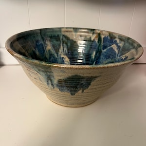 Könnte beinhalten: Eine handgefertigte Keramikschale mit einem breiten, offenen Design. Die Schale hat eine beige Außenseite mit blauen und grünen Akzenten im Inneren und am Rand. Die Schale hat eine strukturierte Oberfläche und eignet sich zum Servieren.