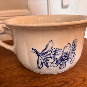 May include: Cream ceramic chamber pot with a blue floral pattern. It features a handle and a wide rim. The floral design is on the front. The pot is on a wooden surface. The pot's dimensions are approximately 20 cm in diameter.