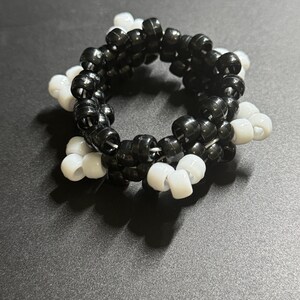 以下が含まれることがあります： 白と黒のビーズブレスレット。ブレスレットは、小さな丸いビーズを繋ぎ合わせて円形に作られています。ビーズは黒と白が交互に配置され、コントラストのある模様を作り出しています。ブレスレットは暗い表面に表示されています。