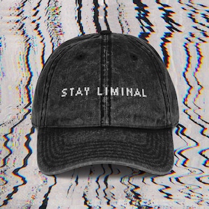 May include: Black baseball cap with the text "STAY LIMINAL" in white pixelated font. The cap has a curved brim and a distressed, vintage look. The background features a distorted, colorful pattern.