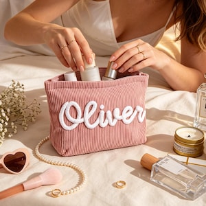 May include: A pink corduroy cosmetic bag with the name "Oliver" in white script. The bag is open, revealing several cosmetic products. Other items include heart-shaped sunglasses, a makeup brush, perfume, a candle, and a white rose.