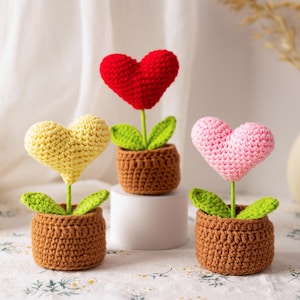 May include: Three crochet heart-shaped flowers in brown pots. One is red, one is yellow, and one is pink. Each has green leaves and a brown pot. The flowers are on a white surface with a white curtain in the background.
