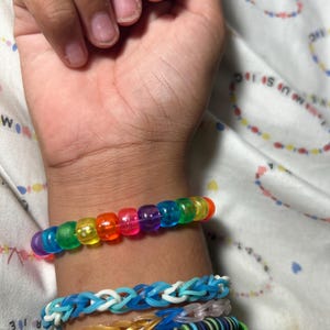 May include: A collection of colorful bracelets adorn a wrist. The top bracelet features translucent, rainbow-colored beads. Below are blue and white woven bracelets, a gold-toned bracelet, and a green bracelet with text.