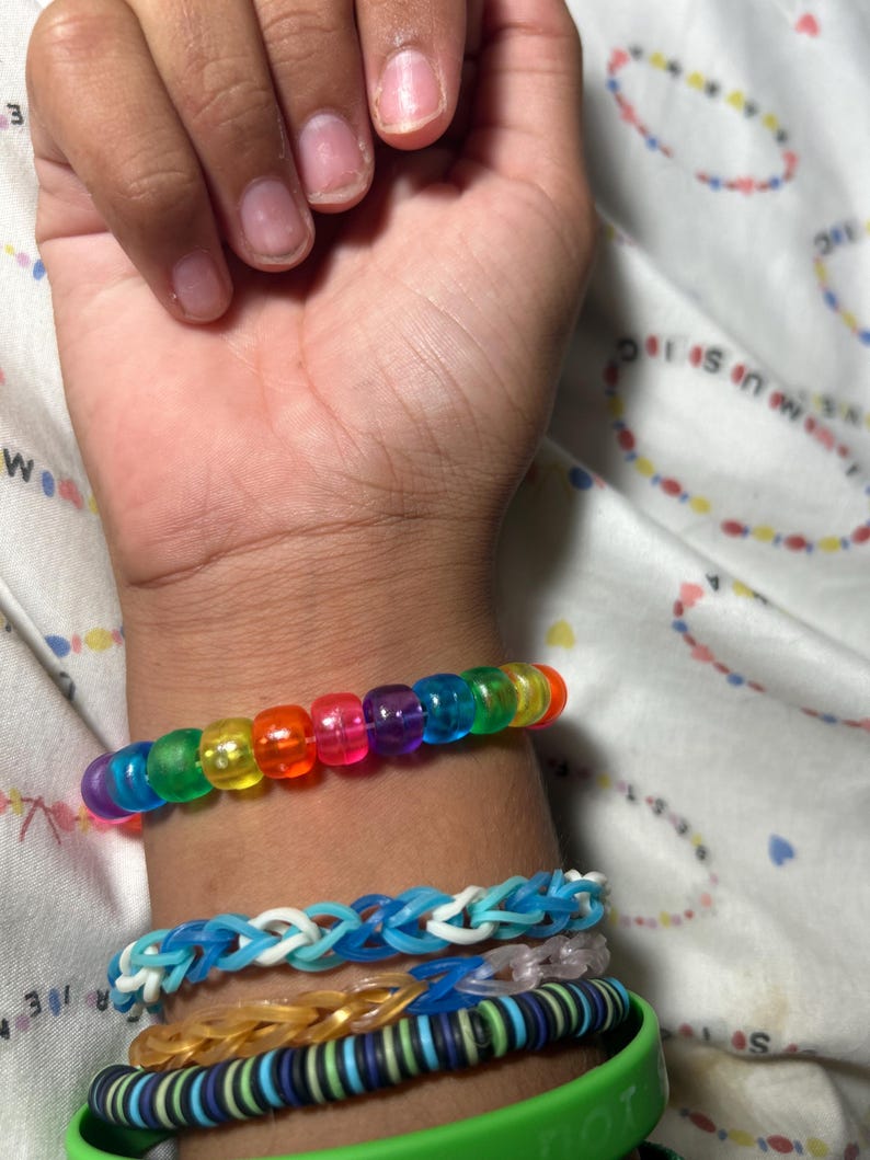 May include: A collection of colorful bracelets adorn a wrist. The top bracelet features translucent, rainbow-colored beads. Below are blue and white woven bracelets, a gold-toned bracelet, and a green bracelet with text.