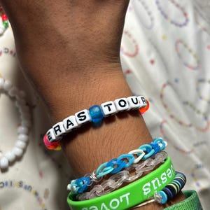 May include: A wrist adorned with multiple bracelets. One bracelet spells "ERAS TOUR" with white letter beads. Other bracelets include a green band with "Jesus Loves" in white, and blue and clear rubber band bracelets.