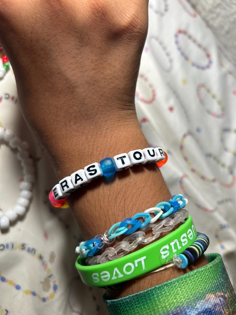 May include: A wrist adorned with multiple bracelets. One bracelet spells "ERAS TOUR" with white letter beads. Other bracelets include a green band with "Jesus Loves" in white, and blue and clear rubber band bracelets.