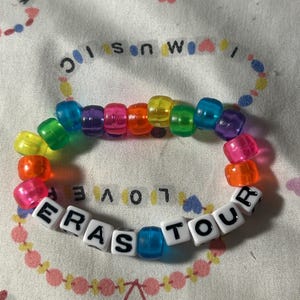 May include: A colorful beaded bracelet with the text "ERAS TOUR" spelled out in white letter beads. The bracelet features a rainbow of translucent beads and is displayed on a patterned fabric.