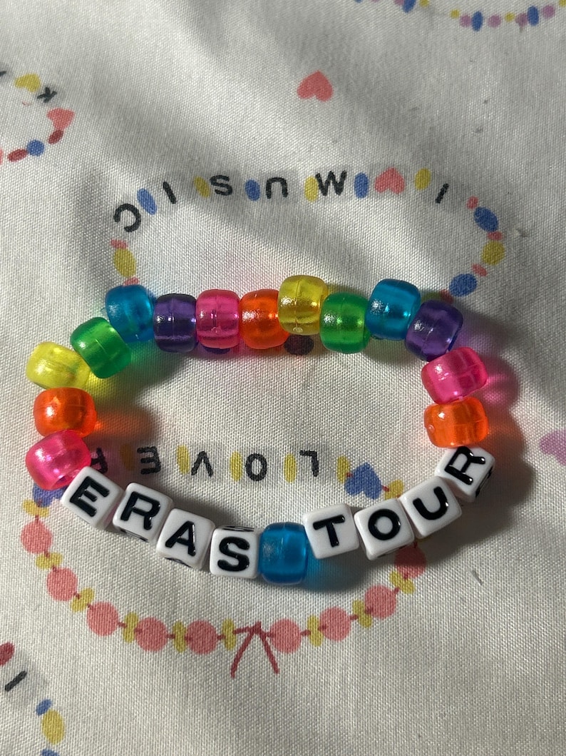 May include: A colorful beaded bracelet with the text "ERAS TOUR" spelled out in white letter beads. The bracelet features a rainbow of translucent beads and is displayed on a patterned fabric.