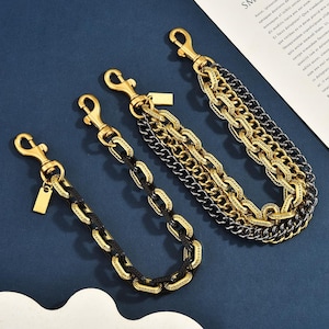 May include: Three metal chains featuring a mix of gold and black links. Each chain is equipped with a gold-toned lobster clasp at one end and a small rectangular metal tag. The chains vary in thickness and link design, showcasing a layered appearance.