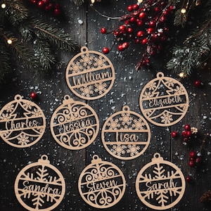 May include: Wooden Christmas ornaments with personalized names, including "William," "Elizabeth," and "Charles." Each ornament features a unique design with snowflakes and a tree. The ornaments are displayed on a dark wooden surface with festive decorations.