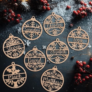 May include: Wooden Christmas ornaments with names like "Sandra", "Charles", and "Lisa" are displayed. Each ornament is circular with a cutout design featuring snowflakes, trees, and other festive elements. The ornaments are made of light brown wood.