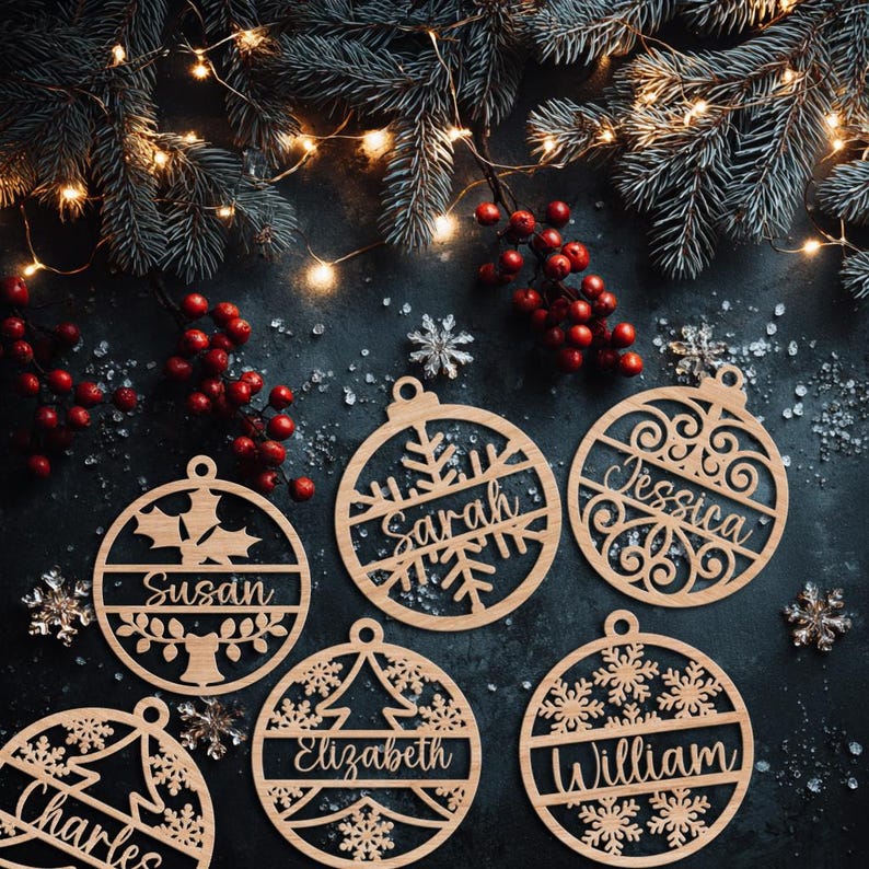 May include: Wooden Christmas ornaments with names like Sarah, Jessica, and William are displayed. The ornaments are round with decorative cutouts of snowflakes, holly leaves, and a Christmas tree. The background includes pine branches, red berries, and fairy lights.