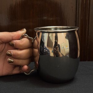 May include: A shiny, stainless steel mug with a curved handle. The mug has a rounded body and a polished, reflective surface. The handle is a simple, elegant design. The mug is held by a hand with glittery nail polish.