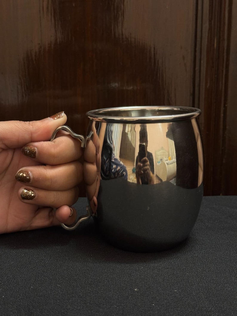 May include: A shiny, stainless steel mug with a curved handle. The mug has a rounded body and a polished, reflective surface. The handle is a simple, elegant design. The mug is held by a hand with glittery nail polish.