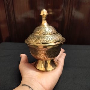 May include: A gold-colored, lidded bowl with intricate floral patterns. The bowl sits on a pedestal base and has a small, rounded handle on the lid. The bowl is held in a hand, and the background is dark.