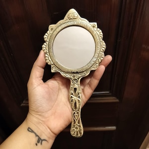 Peut inclure: Un miroir de poche de style vintage avec un cadre et une poignée dorés ornés. Le miroir rond est entouré d'un bord décoratif festonné. La poignée présente un motif floral complexe.