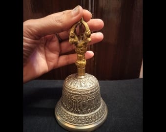 May include: An ornate silver-colored bell with a detailed floral pattern and a gold-colored handle. The handle features a decorative design. The bell is held by a hand, showcasing its size and craftsmanship. The bell is likely used for ceremonial or decorative purposes.