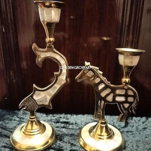 Peut inclure: Deux bougeoirs décoratifs en laiton. L'un présente un motif de poisson, l'autre un cheval. Les deux ont des incrustations blanches et reposent sur une base ovale. Les bougeoirs ont une finition dorée et sont posés sur un fond sombre.