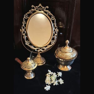 May include: A gold-toned vanity mirror with an oval frame and ornate floral detailing, accompanied by two decorative incense burners and a lidded bowl. A small cluster of white flowers adds a delicate touch to the arrangement.