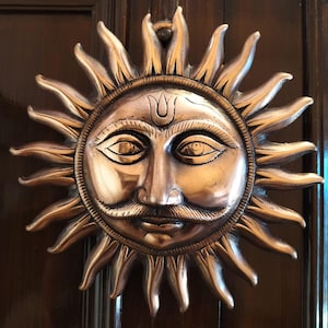 Op de afbeelding: Een bronzen deurklopper met een zonnegelaat en gedetailleerd ontwerp. De zon heeft een snor, gesloten ogen en een decoratief merkteken op het voorhoofd. De zonnestralen stralen naar buiten. De klopper is gemonteerd op een donkerbruine houten deur.