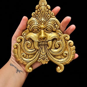 May include: A gold-coloured metal wall plaque featuring a detailed relief of a mythical creature face with prominent fangs, wide eyes, and intricate swirling scrollwork patterns. The decorative piece has a pointed top and a symmetrical design.