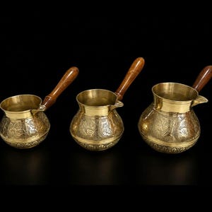 May include: Three brass coffee pots of varying sizes, each featuring a rounded body with intricate embossed floral patterns, a wide pouring spout, and a long, polished wooden handle attached to the side of the metallic vessel.