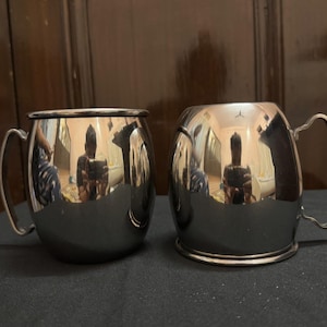 May include: Two stainless steel Moscow Mule mugs with curved handles. The mugs have a shiny, reflective surface, and the interior is not visible. The mugs are sitting on a dark surface.