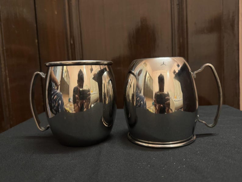May include: Two stainless steel Moscow Mule mugs with curved handles. The mugs have a shiny, reflective surface, and the interior is not visible. The mugs are sitting on a dark surface.
