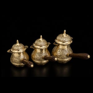 May include: A set of three graduated brass coffee pots featuring intricate floral engravings on the bodies and lids. Each pot has a polished gold-tone finish, a pointed finial on the lid, a small pouring spout, and a dark brown wooden handle.