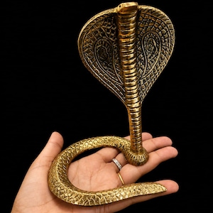 May include: A metallic gold-colored cobra figurine with a raised hood and a coiled body. The surface features intricate, textured scale patterns across the entire snake, with detailed markings visible on the expanded hood area.