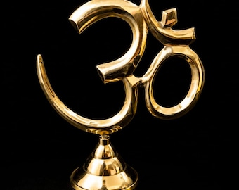 May include: A polished gold-colored metal Om symbol sculpture mounted on a tiered circular base. The Om sign features a smooth, reflective surface with a curved, flowing design that stands upright, showcasing a metallic finish against a dark background.