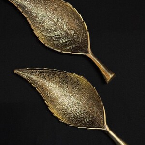 May include: Two gold-coloured leaf-shaped decorative trays. The trays have intricate embossed detailing, resembling the veins of a leaf. Each tray has a stem-like handle. The background is black.