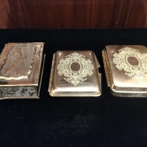 Puede incluir: Tres cajas metálicas antiguas con tapas con bisagras. La caja de la izquierda tiene forma de libro con un diseño floral. Las otras dos cajas son rectangulares con patrones simétricos ornamentados. Todas las cajas tienen un acabado dorado.