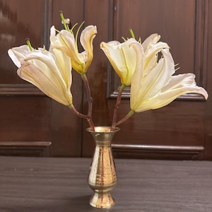 Pode incluir: Um pequeno vaso de latão ornamentado contém várias flores de lírio creme. O vaso tem um design gravado detalhado e está sobre uma superfície de madeira escura. Os lírios têm centros amarelos e estão em plena floração.