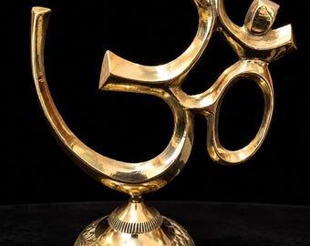 Könnte beinhalten: Eine Skulptur des Om-Symbols aus poliertem Messing auf einem runden Sockel. Der Sockel ist mit detaillierten Blumengravuren und einem strukturierten Rand versehen. Das Om-Zeichen hat eine glatte, reflektierende Oberfläche mit geschwungenen Linien.