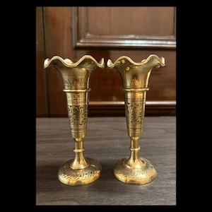 May include: A pair of antique brass vases, featuring ruffled edges and detailed floral patterns. The vases have a slender, elongated form, resting on a circular base. The brass has a warm, golden tone, and they are displayed on a dark wooden surface.