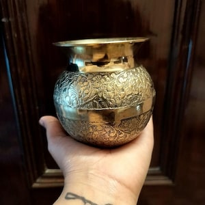 以下が含まれることがあります： 精巧な花柄と広い縁が付いた、小さくて丸い真鍮製のポット。 ポットの中央にはバンドがあり、手で持っています。 背景は暗い木製のドアです。