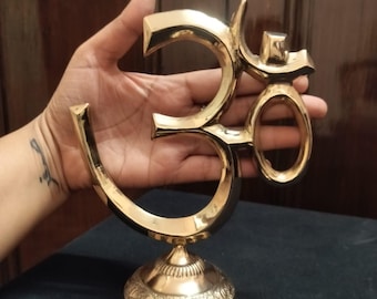May include: A polished gold-toned metal Om symbol sculpture featuring a smooth, reflective surface. The Om sign is mounted on a circular base with intricate, embossed floral and geometric patterns around the lower edge of the stand.