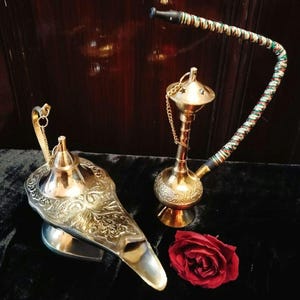 May include: An ornate brass genie lamp and hookah are displayed. The lamp is silver with intricate floral designs. The hookah has a decorative base, a chain, and a flexible hose with a colorful pattern. A red rose is also present.
