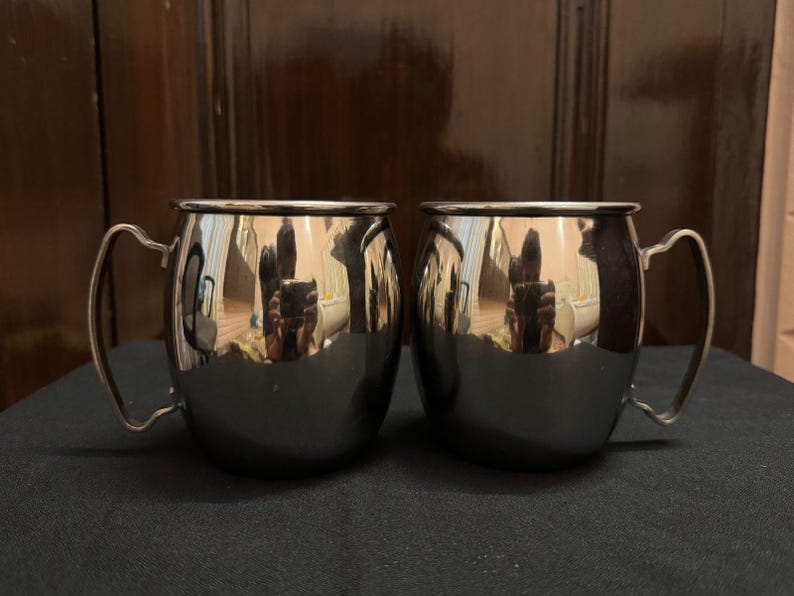 May include: Two stainless steel mugs with curved handles. The mugs have a shiny, reflective surface, and a rounded body. The mugs are sitting on a dark surface, and the background is a dark brown.