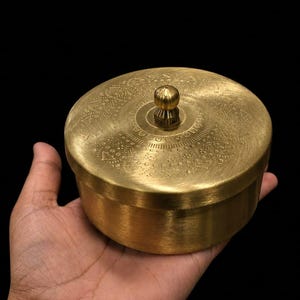 Op de afbeelding: Een ronde messing container met een afneembaar deksel voorzien van een ingewikkeld, geëtst geometrisch patroon en een kleine, centrale knopgreep. Het metalen oppervlak heeft een geborstelde, gouden afwerking tegen een zwarte achtergrond.