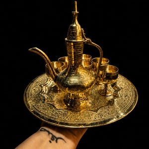 May include: A polished gold-colored metal coffee set consisting of a tall, slender dallah coffee pot with a long curved spout and a matching handle, accompanied by six small stemmed cups, all arranged on a round, intricately engraved serving tray.