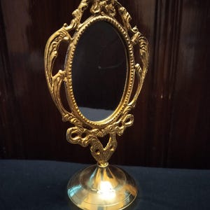 May include: A small, ornate, gold-colored vanity mirror. The oval mirror is framed by an elaborate, decorative border. The mirror sits on a round, gold-colored base. The overall design is vintage and elegant.