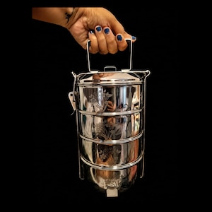 May include: A four-tier stainless steel tiffin carrier featuring a polished reflective finish and delicate floral engravings on each cylindrical container. The stackable food container includes a secure locking mechanism and a sturdy metal handle.