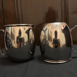May include: Two stainless steel mugs with curved handles. The mugs have a shiny, reflective surface, and the interior is not visible. The mugs are sitting on a dark surface.