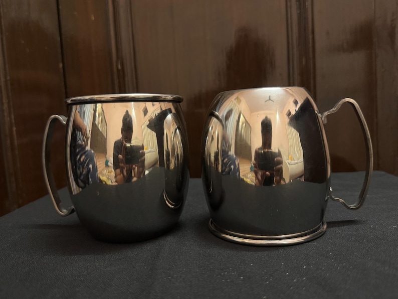May include: Two stainless steel mugs with curved handles. The mugs have a shiny, reflective surface, and the interior is not visible. The mugs are sitting on a dark surface.