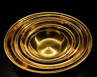 Puede incluir: Un conjunto de cuencos circulares anidados hechos de metal pulido de color dorado. Los cuencos están dispuestos uno dentro del otro, creando una serie de anillos concéntricos. La superficie metálica refleja la luz y resalta los bordes.
