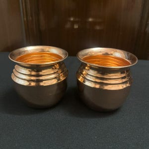 May include: Two small, copper-colored pots with a rounded base and a wide, open top. The pots have a series of horizontal ridges around the body and a shiny, reflective interior. The pots are displayed on a dark surface.