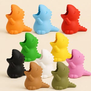 Könnte beinhalten: Eine Sammlung von zehn kleinen, dinosaurierförmigen Silikonhaltern in verschiedenen Unifarben wie Orange, Hellblau, Rot, Grün, Gelb, Schwarz, Weiß, Braun, Olivgrün und Pink. Jede Figur verfügt über ein geöffnetes Maul und eine glatte Oberfläche.