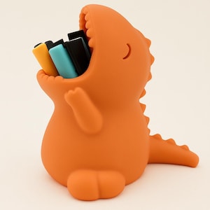 Könnte beinhalten: Orangefarbener Schreibtisch-Organizer in Dinosaurierform, der farbige Stifte enthält. Der Organizer hat ein großes offenes Maul mit Zähnen, kleine Arme und einen Schwanz. Die Stifte sind gelb, blau und schwarz.